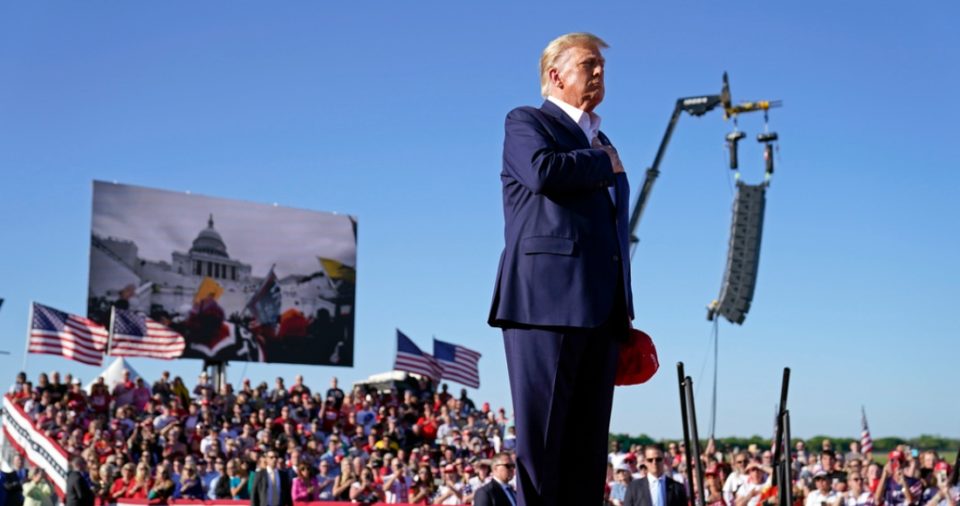 El expresidente Donald Trump escucha de manera solemne la canción "Justice for All" (Justicia para todos) durante un acto político en el aeropuerto regional de Waco, Texas, el sábado 25 de marzo de 2023, mientras una pantalla gigante al fondo difunde imágenes del asalto al Capitolio en 2021.